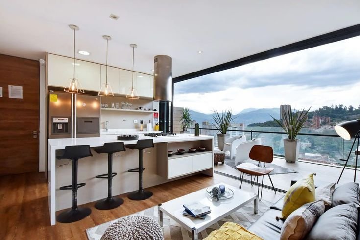 Modern open-plan Airbnb apartment with a bright kitchen, bar stools, cozy living area, and floor-to-ceiling windows overlooking a mountain cityscape.