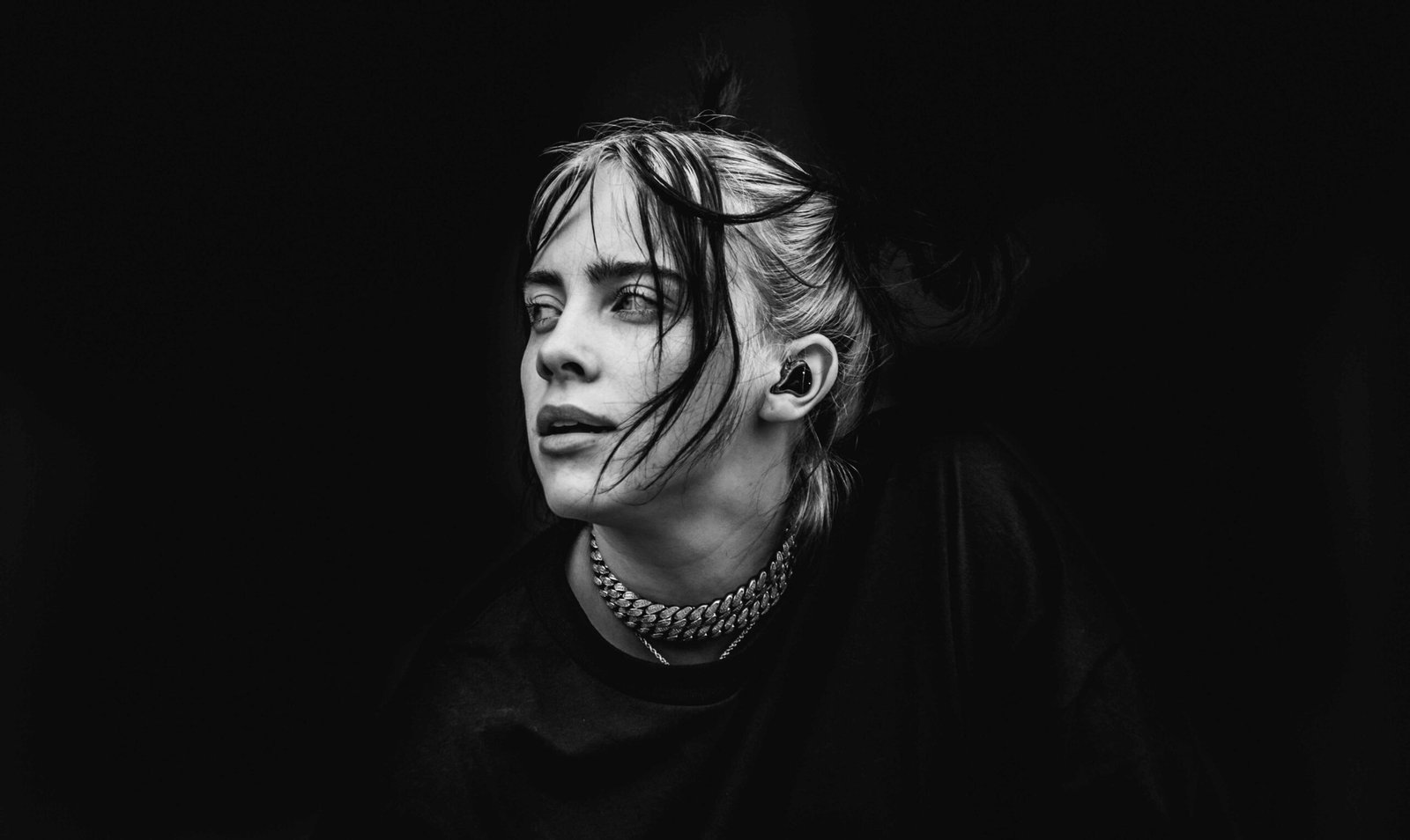 Billie Eilish performing on stage, shown in a black-and-white close-up portrait with windblown hair, wearing a chain necklace and in-ear monitor against a dark background.