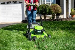 Person mowing a residential lawn with a green electric lawn mower in front of a house.