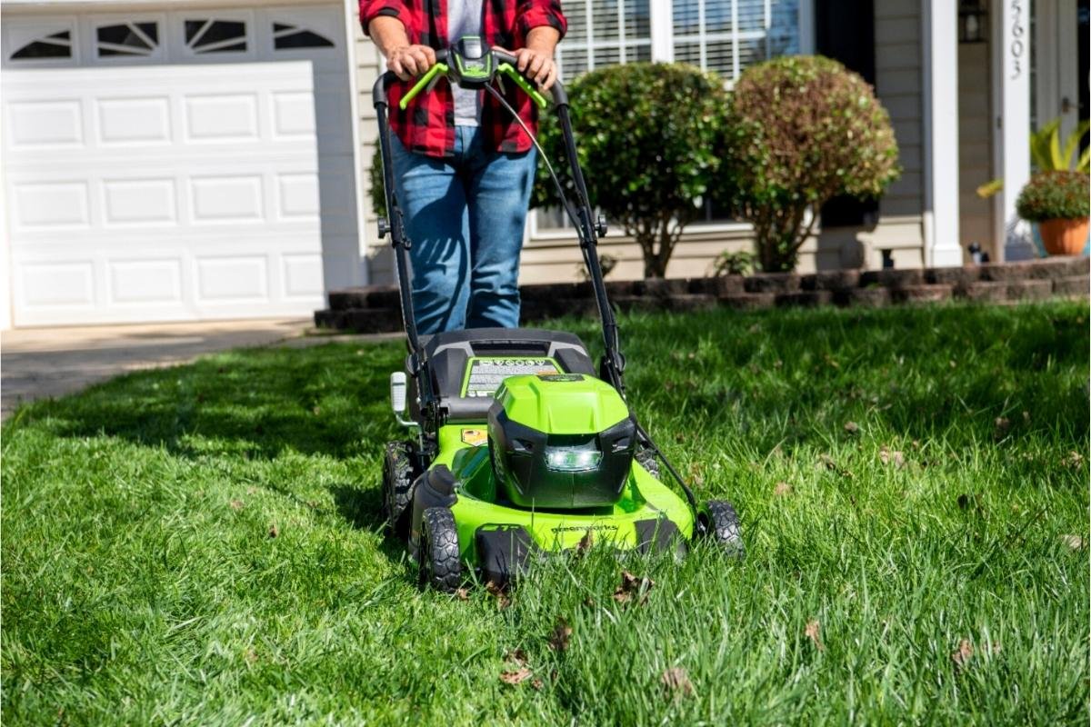 Person mowing a residential lawn with a green electric lawn mower in front of a house.