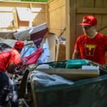 Two junk removal workers in red uniforms clearing a large pile of debris and cardboard boxes in an outdoor enclosure.