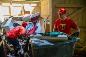 Two junk removal workers in red uniforms clearing a large pile of debris and cardboard boxes in an outdoor enclosure.