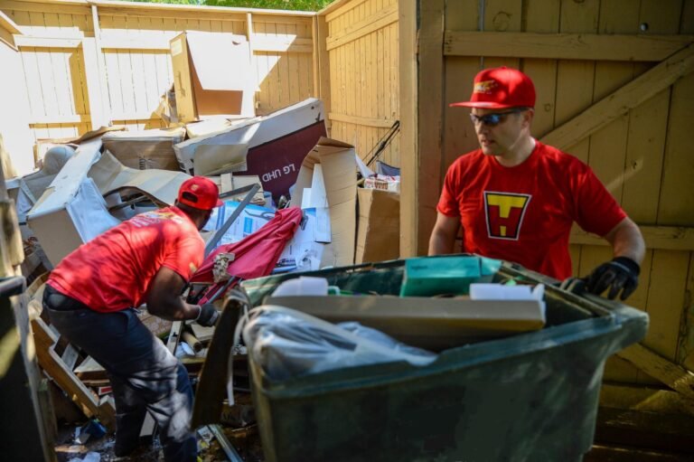 Two junk removal workers in red uniforms clearing a large pile of debris and cardboard boxes in an outdoor enclosure.