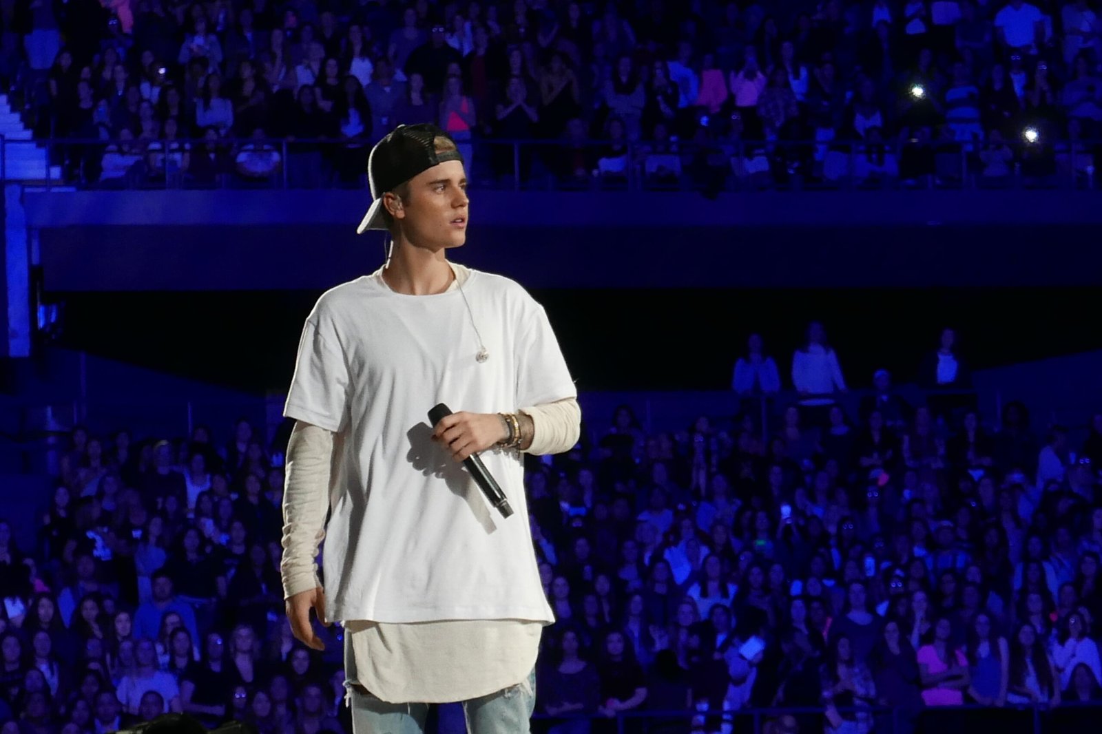 Justin Bieber wearing a white shirt and holding a microphone stands onstage in front of a large concert crowd, featured in an article about Justin Bieber net worth.