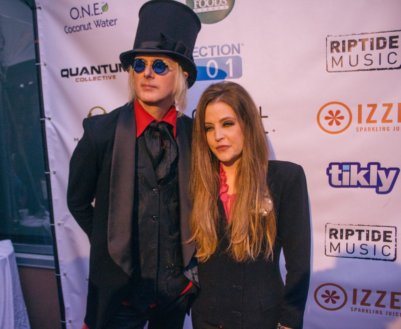 Lisa Marie Presley and men dressed in formal clothing posing on a red carpet in front of event sponsor logos, featured in an article about Lisa Marie Presley net worth.
