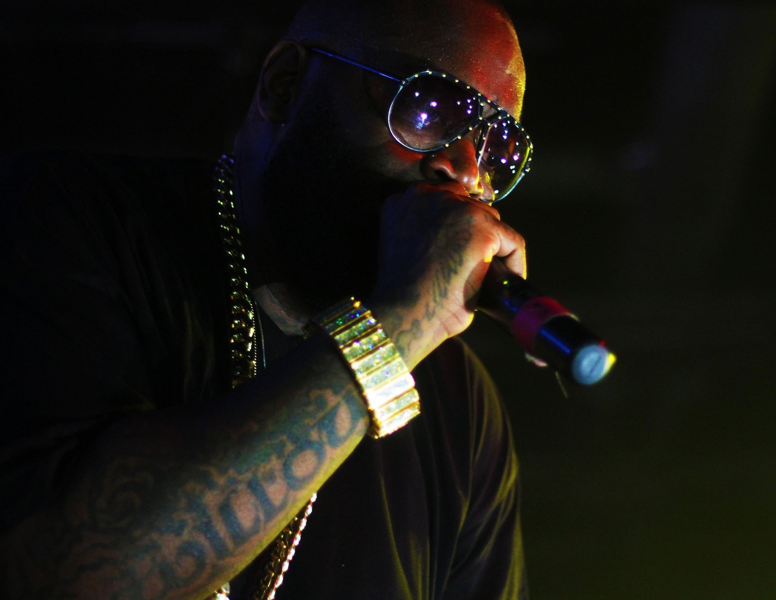 Rick Ross wearing sunglasses and gold jewelry raps into a microphone under stage lighting, featured in an article about Rick Ross net worth.