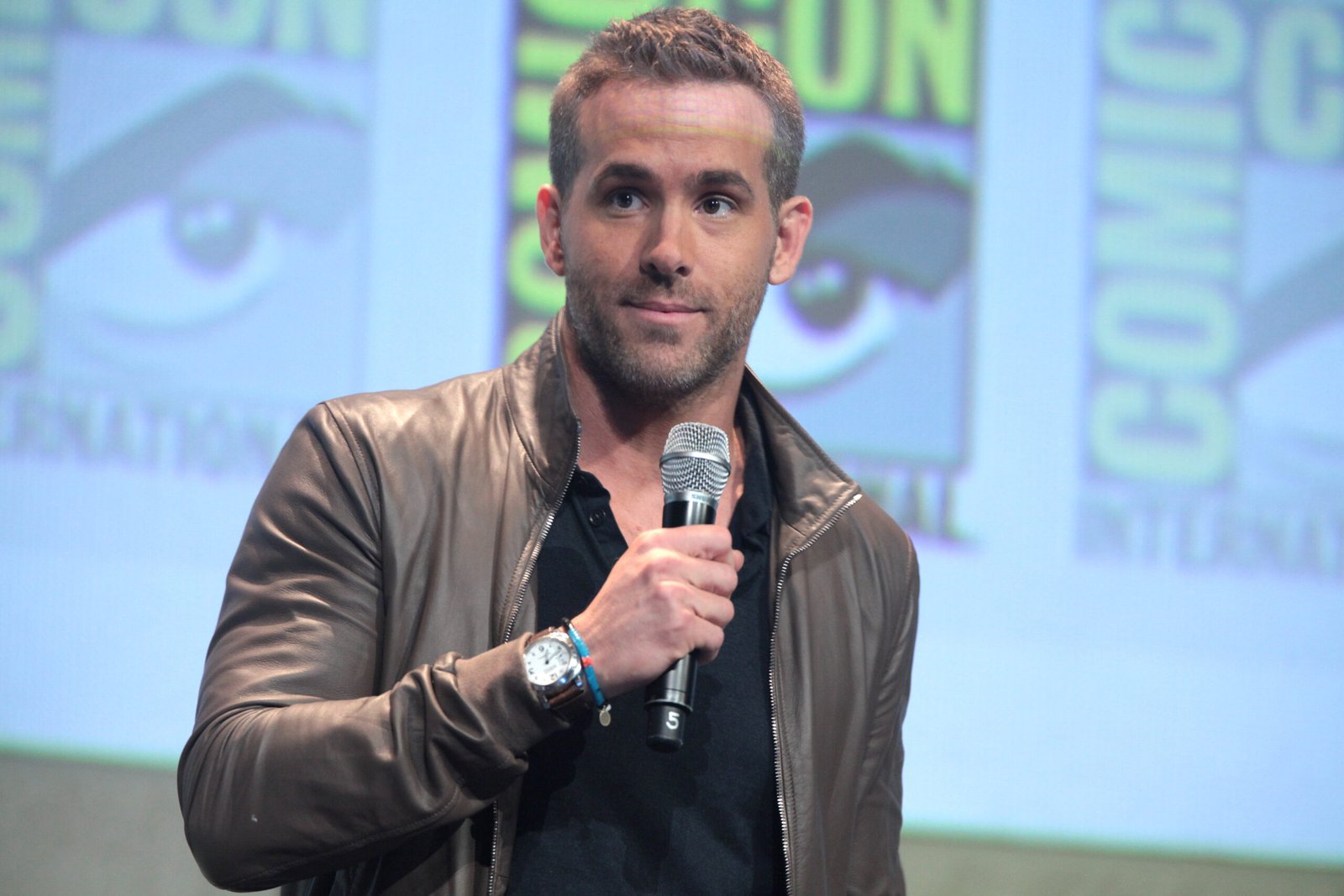 Ryan Reynolds holding a microphone while speaking on stage at a comic convention, featured in an article about Ryan Reynolds net worth.
