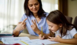 A math tutor helps a young student solve a problem, using her fingers to demonstrate a counting strategy while the child writes answers in a workbook.