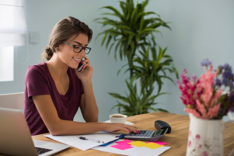 A small business owner sits at a desk using a calculator while talking on the phone, surrounded by paperwork, a laptop, and colorful notes, representing bookkeeping and financial management.