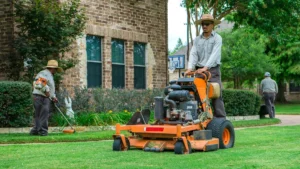 A lawn care crew working on a residential property, with one worker mowing the grass using a commercial mower and another trimming edges near the home’s landscaping.