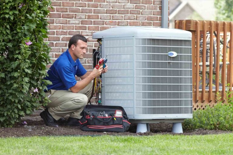 A technician kneels beside an outdoor air conditioning unit, using HVAC gauges and tools to check the system while a tool bag rests on the ground nearby.