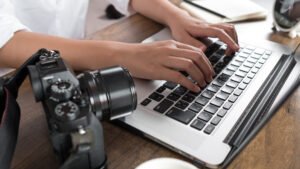Photographer editing on a laptop with a digital camera beside them on a wooden desk.
